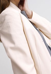 Blazer texturizado de color rosa claro con un corte entallado, frente abierto y hombros ligeramente acolchados, superpuesto sobre una prenda gris.