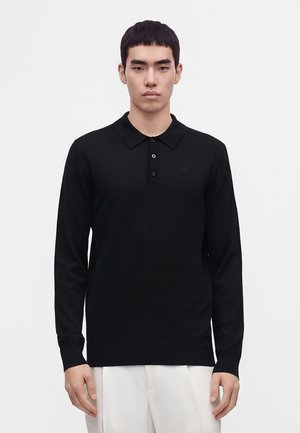 Junger Mann trägt ein schwarzes langärmeliges Poloshirt und weiße Hose und steht vor einem einfachen hellen Hintergrund.