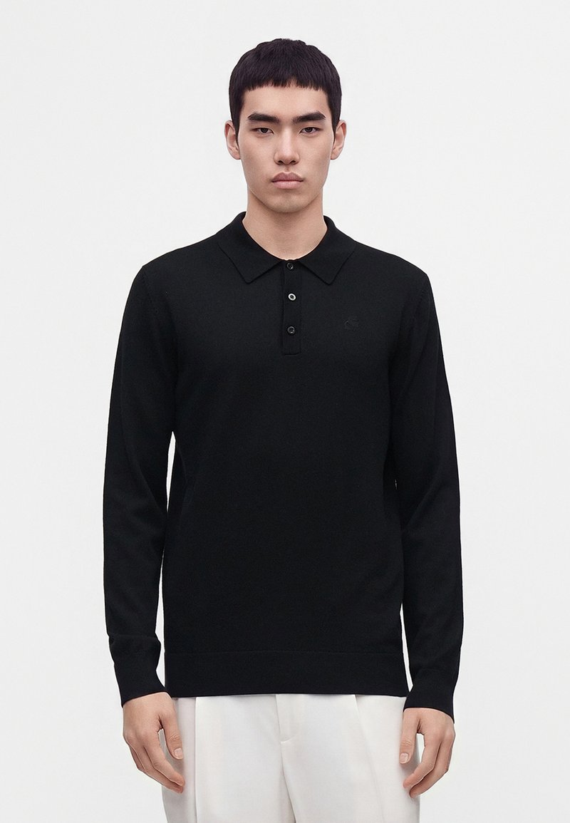 Jeune homme portant un polo noir à manches longues et un pantalon blanc, debout sur un fond clair uni.
