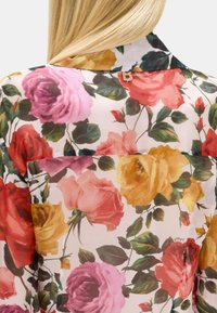 Camicia con motivo floreale caratterizzata da grandi rose colorate in rosa, giallo e rosso su uno sfondo chiaro. Presenta un piccolo badge metallico a forma di 'B' sul collo.