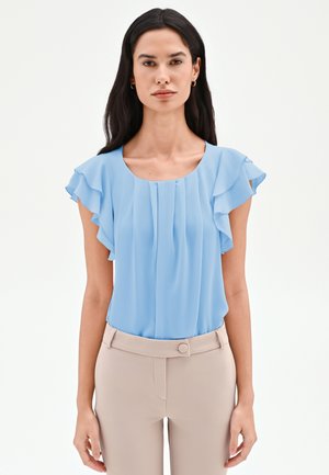 Donna con capelli lunghi e scuri che indossa una camicetta a maniche corte azzurra con volant infilata in pantaloni beige a vita alta, su uno sfondo semplice.