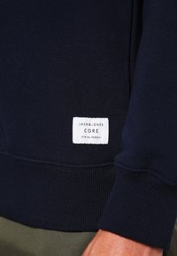 Sudadera azul marino con puños y dobladillo acanalados, que presenta una etiqueta blanca con el texto "JACK & JONES CORE" y un número de etiqueta. Material de mezcla de algodón.