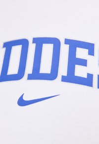 Bílý tričko s velkým modrým nápisem "GODDESS." Obsahuje modré logo Nike, hladkou strukturu a standardní design s kulatým výstřihem.