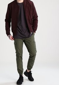 Burgundy bomberjacka med ribbat krage, svart t-shirt, olivgröna cargobyxor med sidofickor och svarta sneakers. Avslappnad streetwear-stil.