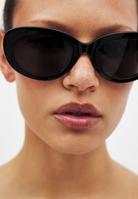 Storebriller i sort med en glat, blank finish og mørke glas. Nærbillede der fremhæver rammes formen og ansigtstræk.