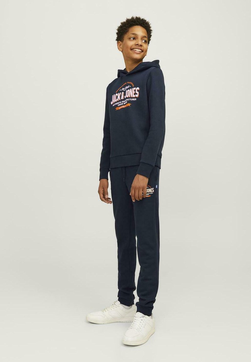 Mørkeblå hoodie og joggersæt lavet af blødt stof. Hoodien har et logo, kængurulomme og ribbede manchetter. Sættes sammen med hvide sneakers.