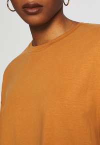 Tričko s krátkým rukávem a kulatým výstřihem v teplé oranžové látce s jemnou texturou a mírnými odstíny, doplněné zlatým kruhovým náušnicem.