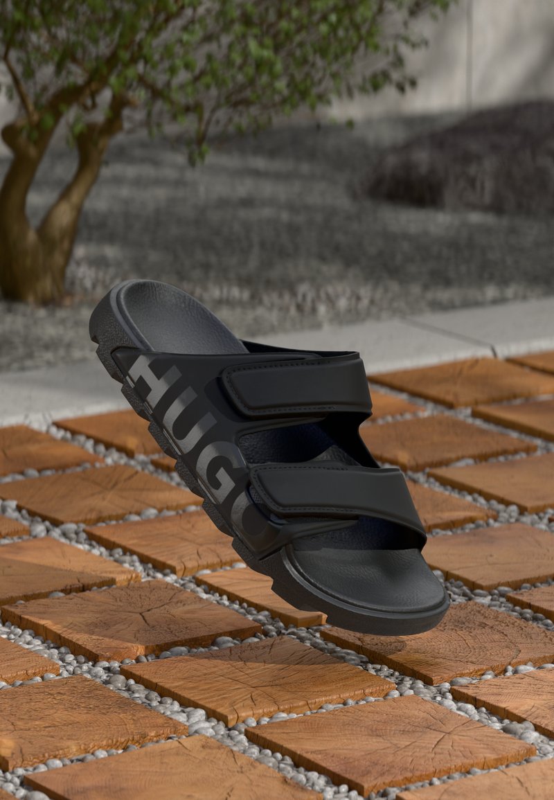 Svart skyvesandal med to brede stropper, med "HUGO"-logo. Glatt tekstur, formet såle, hvilende på en trefliser med grusdetaljer.
