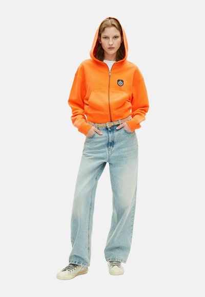 Oranje hoodie met rits en een gecentreerd embleem, gecombineerd met lichtblauwe wide-leg jeans en beige sneakers met grijze veters.