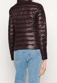 Giubbotto imbottito color bordeaux con design trapuntato orizzontale, finitura lucida, collo alto e silhouette aderente. Abbinato a jeans azzurri.