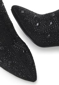Tacones negros de punta afilada con tejido texturizado, adornados con rhinestones negros y tachuelas en forma de pirámide, que cuentan con una suela suave y una silueta esbelta.