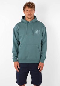 Sweat à capuche teal avec poche frontale, cordons ajustables et motif circulaire sur la poitrine gauche. Associé à un short navy. En mélange de coton.