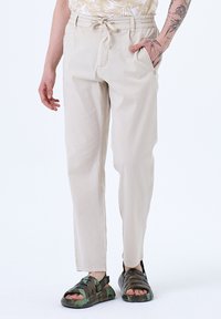 Pantalons en coton beige avec une coupe décontractée, taille ajustable et poches latérales, associés à des sandales en camouflage foncé.