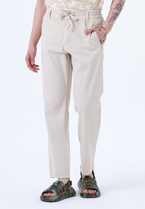 Beige katoenen broek met een relaxte pasvorm, verstelbare tailleband en zijzakken, gecombineerd met donkere camouflage sandalen.