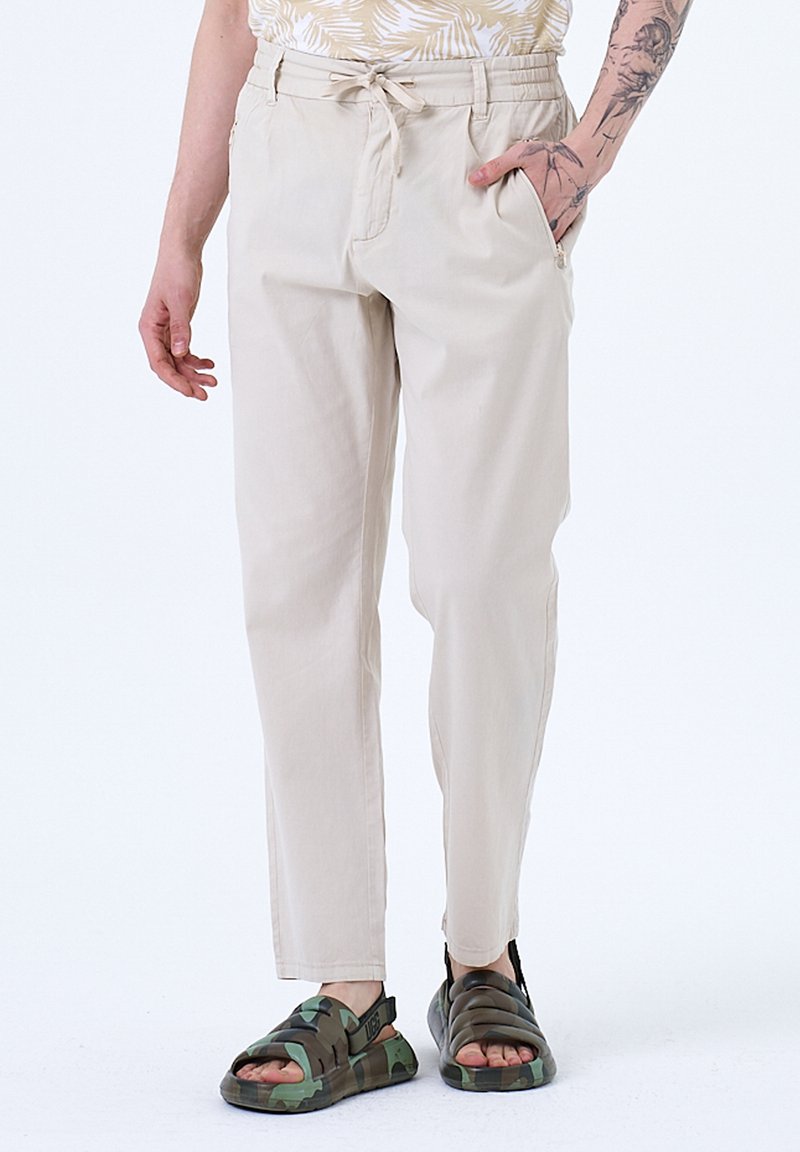 Pantalons en coton beige avec une coupe décontractée, taille ajustable et poches latérales, associés à des sandales en camouflage foncé.