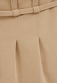 Beige tyg med en slät textur som har plisseringar och en matchande bältdetalj. Sytt accentuerar den strukturerade designen.