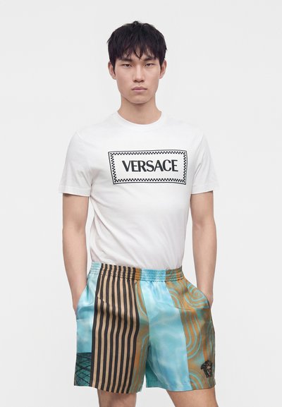 Jeune homme portant un t-shirt blanc Versace et un short rayé et à motifs coloré, les mains dans les poches.