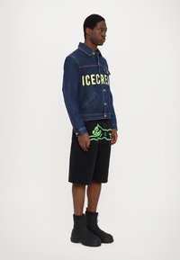Denimjakke med "ICECREAM" trykket i gult, sammen med svarte shorts med et grønt grafisk design, og svarte glidelås-støvler.