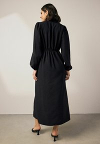 Czarna midi sukienka z teksturowanej tkaniny, elastyczną talią i bufiastymi rękawami. Stylizowana z odkrytymi czubkami butów na obcasie, prezentująca smukłą sylwetkę.