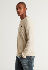 Beige strukturierte Langarmbluse mit Rundhalsausschnitt und kontrastierenden Schulterakzenten, versehen mit einem kleinen schwarzen Logo auf der Brust.
