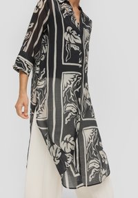 Durchsichtige schwarze Bluse mit einem Blatt- und abstrahierten Design in Creme, Knopfverschluß vorne, Kragen und hochgekrempelten Ärmeln; seitliche Schlitze für Bewegungsfreiheit.