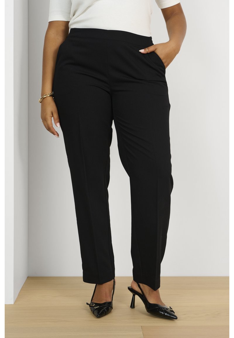 Femme portant un pantalon noir sur mesure et des chaussures slingback noires à talons hauts, debout sur un sol en bois clair contre un mur blanc.