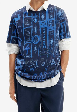 Homme portant un haut à manches courtes bleu marine avec des motifs bleus abstraits et le texte "Desigual" sur une chemise blanche à manches longues, pantalon bleu marine, mains dans les poches.