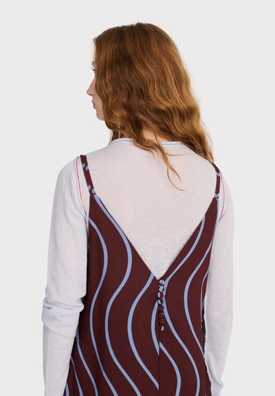 Femme aux longs cheveux roux ondulés portant un haut blanc à manches longues sous une robe marron avec des lignes ondulées bleues et des boutons dans le dos.