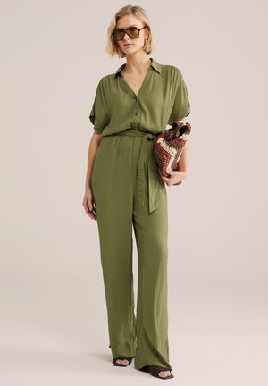 Vrouw in een olijfgroene jumpsuit met een textuur en een tailleband, oversized zonnebril, een meerkleurige geweven clutch in de hand en open schoenen aan.
