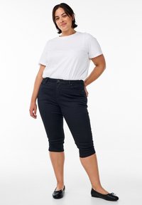 Naine seisab käsi puusal, tal on seljas valge t-särk, mustad poolpüksid ja mustad baleriinikingad valge tausta ees.