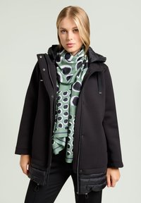 Chaqueta negra con capucha de tela texturizada, con un panel inferior acolchado, combinada con una bufanda verde que muestra patrones circulares en blanco y negro.