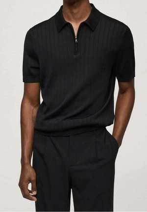 Mand iført en kortærmet sort ribstrikket poloshirt med lynlås foran, kombineret med sorte bukser, stående mod en ensfarvet baggrund.