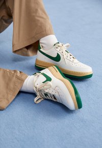 Weiße Nike-Sneaker mit grünen Akzenten, ausgestattet mit einer perforierten Zehenbox, beigem Zwischensohle und strukturiertem grünen Außensohle, kombiniert mit beigen Hosen.