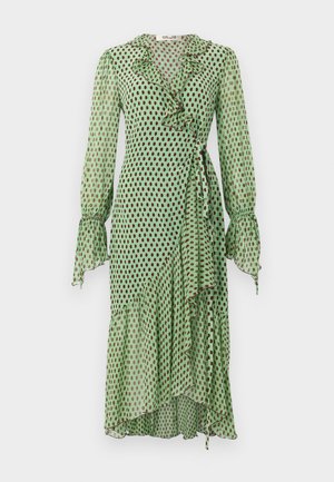 Γυναίκα που στέκεται φορώντας ένα διάφανο πράσινο μίντι φόρεμα τύπου wrap με μαύρα πουά, γιακά με βολάν, μακριά μανίκια και λευκά πέδιλα με λουράκι στον αστράγαλο.