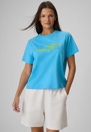 Kobieta o długich brązowych włosach nosi niebieski T-shirt z napisem "Feeling Tropical" i białe szorty, stoi na tle jednolitego szarego tła.