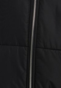 Juodas pūkuotas švarkas su siuvinėjimu, turintis išryškintą sidabrinę užtrauktuko uždarymo sistemą vienoje pusėje. Lygus audinys su minimaliais dizaino elementais.