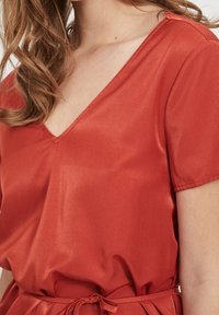 Blouse en satin rouge avec un col en V, manches courtes et ceinture à nouer. Texture lisse avec un léger éclat, présentant une coupe décontractée.