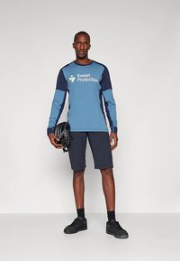 Μακρυμάνικο αθλητικό μπλουζάκι σε μπλε και ναυτικό χρώμα με λογότυπο, συνδυασμένο με ναυτικό σορτς και μαύρα παπούτσια, κρατώντας μαύρο κράνος.