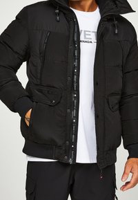 Sort pufferjakke med lynlås og knapper foran, to forlommer og hætte. Har struktureret stof og dekorative detaljer.