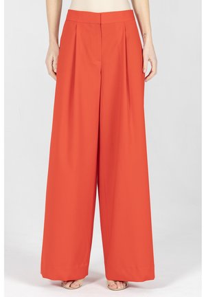 Pomarańczowe spodnie o szerokich nogawkach i wysokim stanie z przednimi zakładkami, noszone przez osobę stojącą prosto, pokazującą stopy w sandałach na otwarte palce.