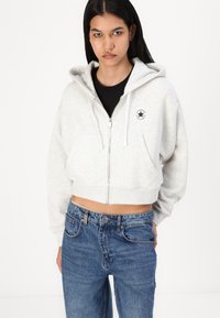 Cropped gray zip-up hoodie made of soft material, featuring a white circular logo and drawstrings, paired with blue denim jeans.