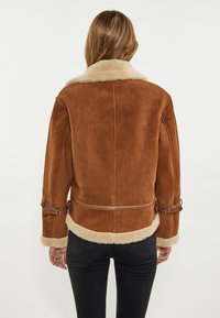 Brun shearlingjakke med en blød beige krave, læderdetaljer, spænde og en tætsiddende design. Glat tekstur og strømlinet form.
