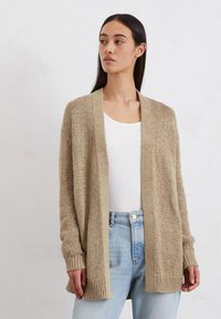Beige gebreide cardigan met open voorkant, lange mouwen en geribbelde manchetten; gedragen over een wit topje en lichtblauwe jeans. Gestructureerde stof.