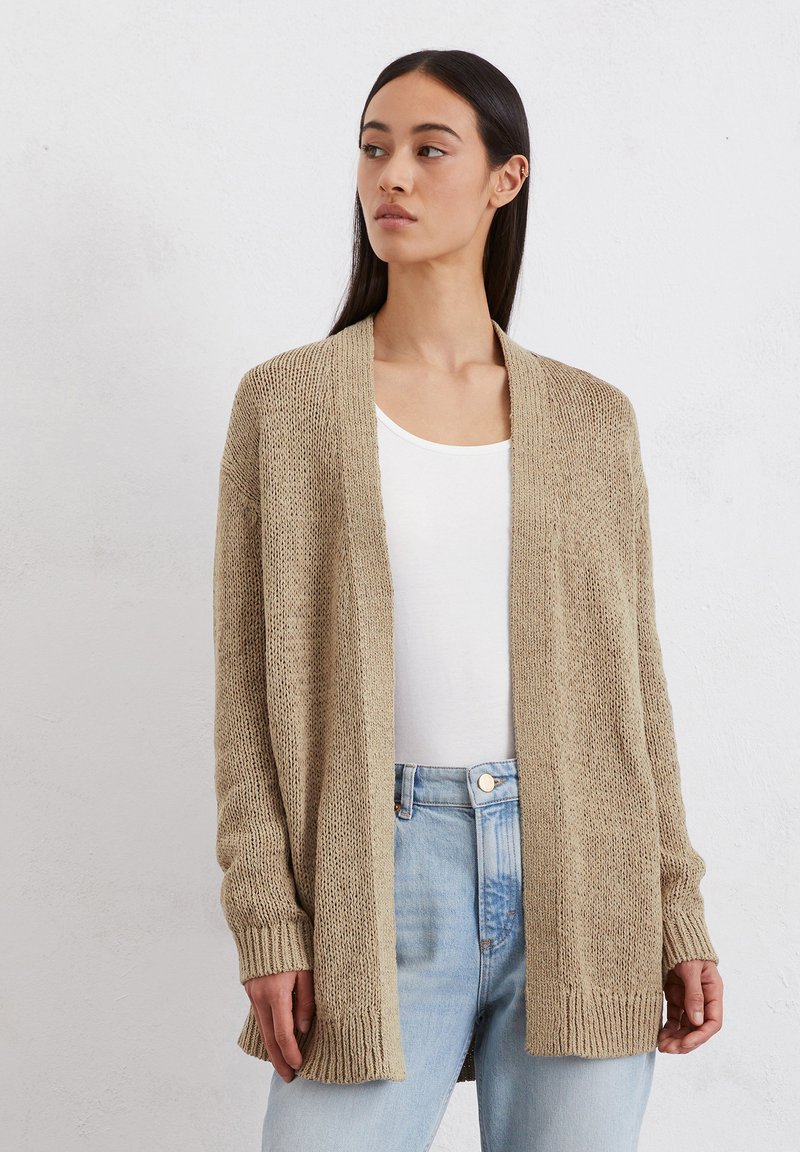 Beige gebreide cardigan met open voorkant, lange mouwen en geribbelde manchetten; gedragen over een wit topje en lichtblauwe jeans. Gestructureerde stof.