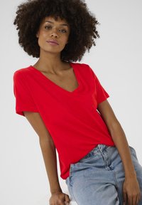 Piros, rövid ujjú póló V-nyakkivágással, puha anyagból készült, laza szabású. Világoskék farmernadrággal párosítva.