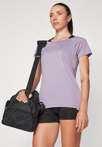 Egy lila Puma pólót és fekete sortot viselő nő, aki egy fekete tornazsákot hord a válla felett, egy egyszínű háttér előtt áll.