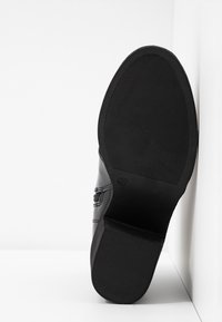 Semelle de chaussure en cuir noir avec une texture antidérapante, inclinée contre une surface d'angle blanche, montrant les détails du talon et de l'avant de la semelle.