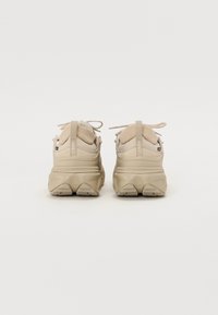 Adidași bej cu un material superior texturat din plasă, guler căptușit și o talpă din cauciuc masivă. Prezintă șireturi și un logo pe călcâi.