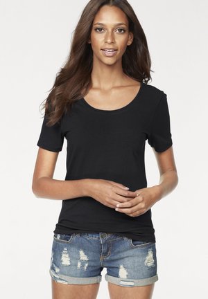Woman with long brown hair wearing a black short-sleeve shirt and ripped denim shorts, standing with hands loosely clasped.