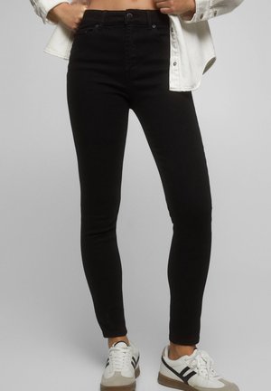 Person wearing black skinny jeans paired with white sneakers and a white jacket, standing against a gray background.